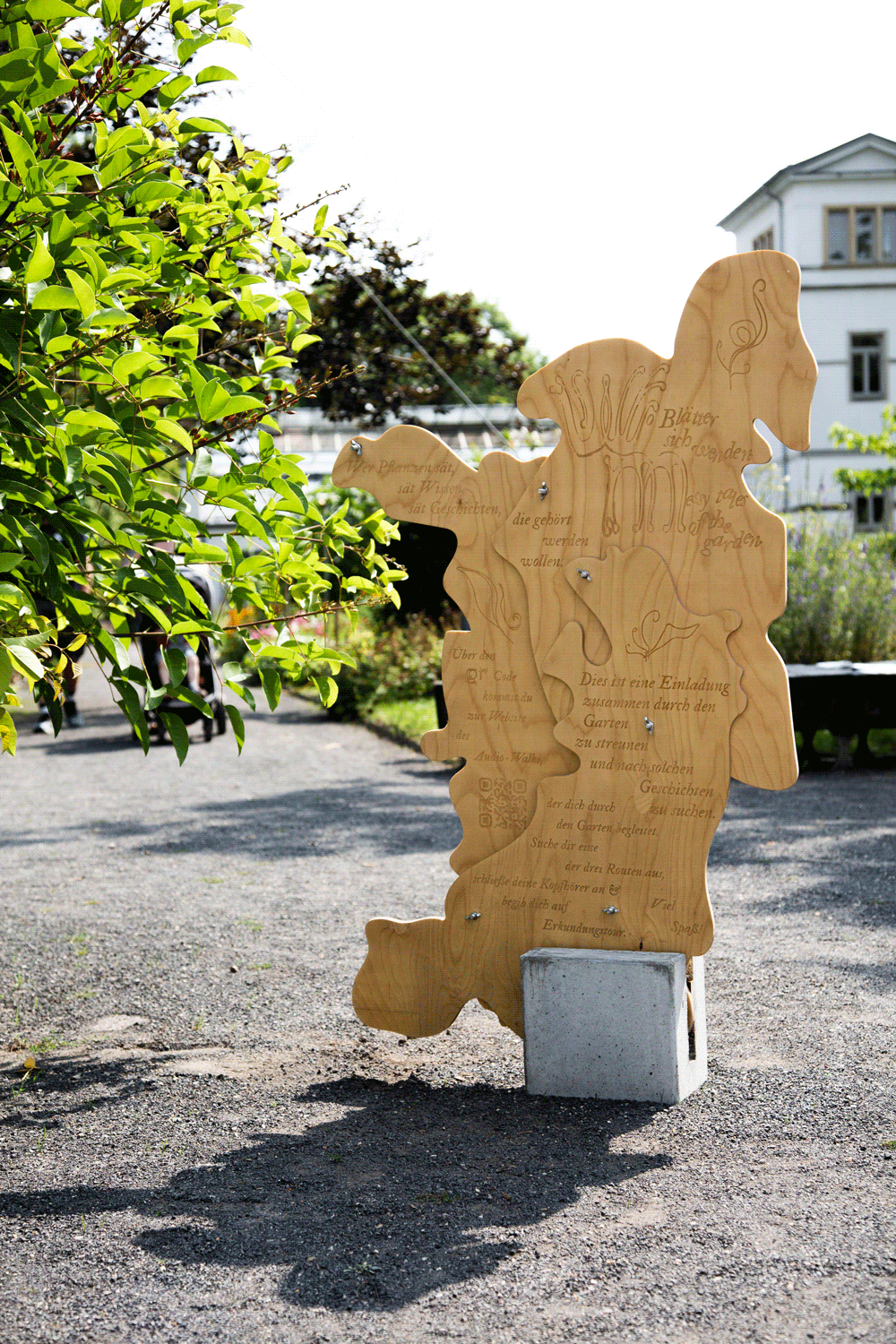 Holzskulptur mit eingravierter Schrift frei stehend mit Pflanzen im Hintergrund